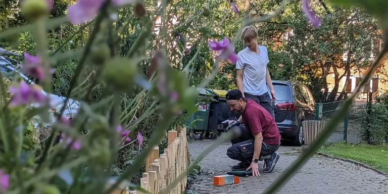 Zwei Personen arbeiten auf einem gepflasterten Gartenweg und bauen einen niedrigen Holzzaun. Im Vordergrund sind unscharfe Blumen zu sehen, im Hintergrund stehen Bäume, Mülltonnen und ein geparktes Auto.