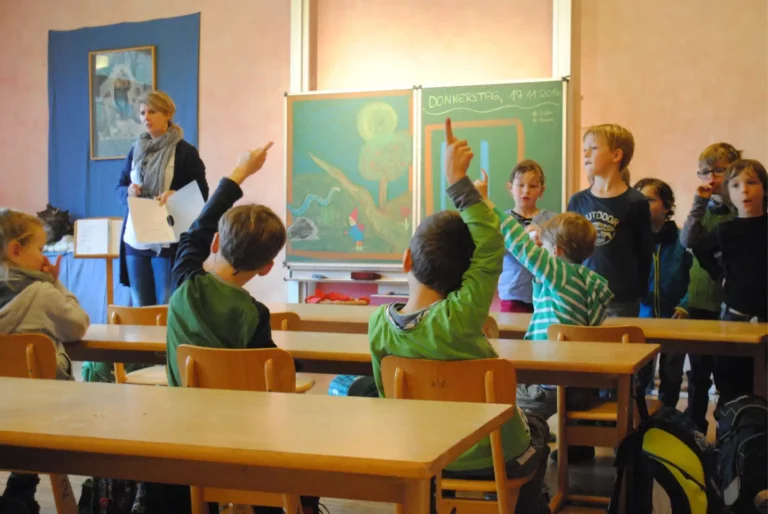 Lehrerin unterrichtet Kinder im Klassenzimmer, mehrere Schülerinnen und Schüler melden sich.