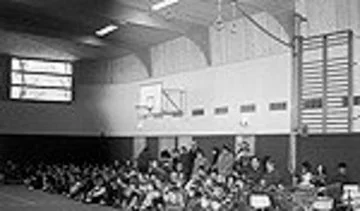 Turnhalle mit vielen sitzenden Kindern und einem Basketballkorb an der Wand.