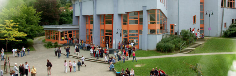 Viele Menschen stehen vor dem Gebäude der Rudolf-Steiner-Schule mit großen orangefarbenen Fenstern und Eingängen.