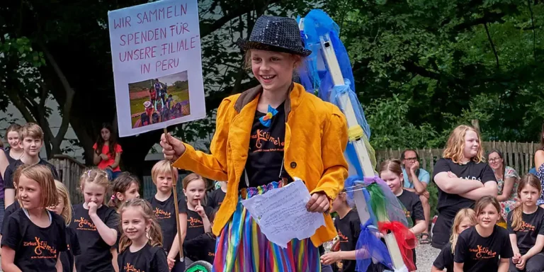 Kind hält ein Schild für eine Spendenaktion, andere Kinder sitzen im Hintergrund.